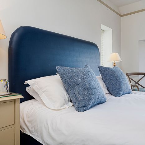 A neatly made bed with a large blue headboard, white bedding, and blue accent pillows in a bright, modern bedroom with bedside lamps and a window letting in natural light.
