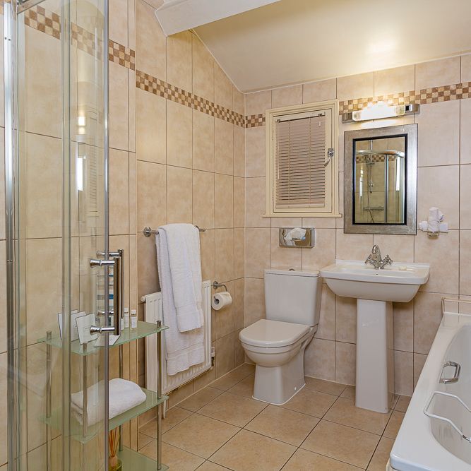 Modern bathroom with beige tiled walls, a glass shower enclosure, a white toilet, pedestal sink, bathtub, and mirror above the sink. Towels and toiletries are neatly arranged.