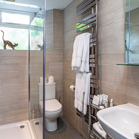 Modern bathroom with glass shower enclosure, toilet, sink, heated towel rack with white towels, and decorative cat figurines on the window.
