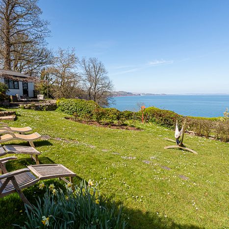 A scenic garden with wooden deck chairs overlooking the ocean, featuring a modern sculpture, green lawn, and a house to the left under clear blue skies.