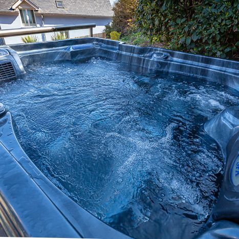 Close-up view of a bubbling hot tub outdoors with bushes and a house in the background.