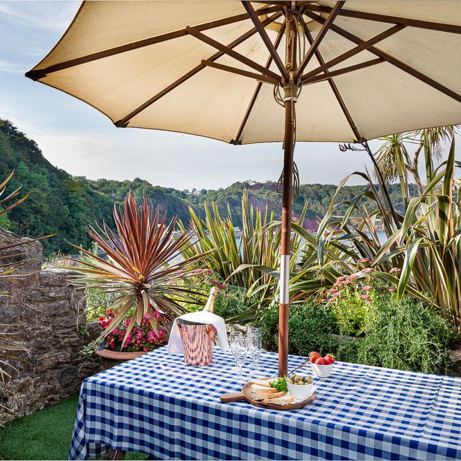 Outdoor picnic table with a blue checkered tablecloth under a large umbrella, surrounded by lush greenery and overlooking a scenic hillside view.