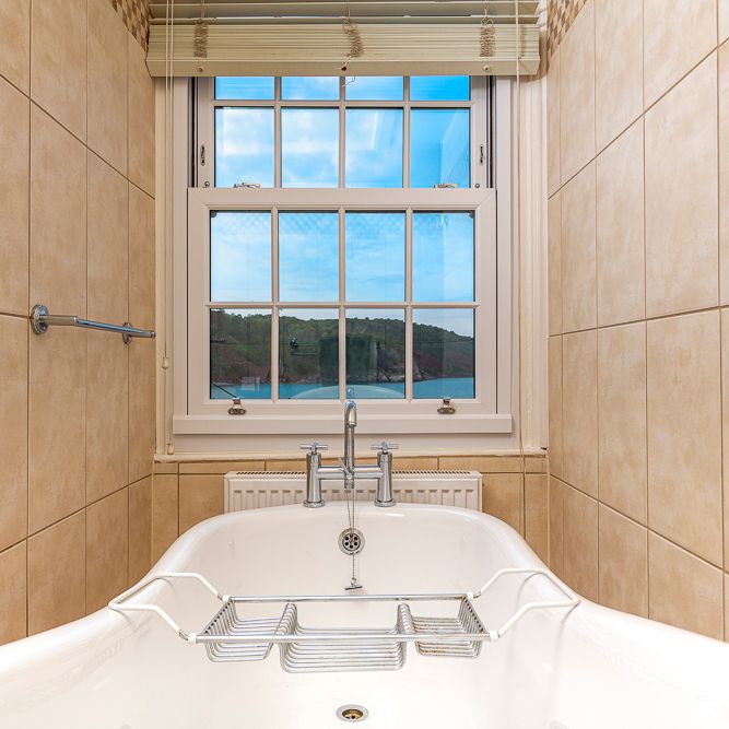 A white bathtub positioned under a large window with tan tiled walls on both sides. The window offers a scenic view of hills, water, and a blue sky outside.