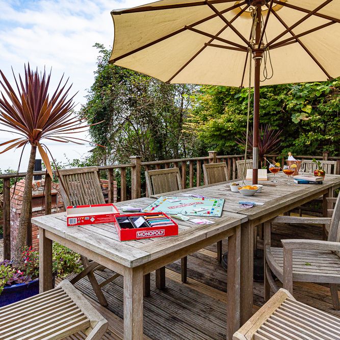 Outdoor wooden patio with a table, chairs, a large umbrella, and a Monopoly board game set up. The patio overlooks water and is surrounded by trees and plants.