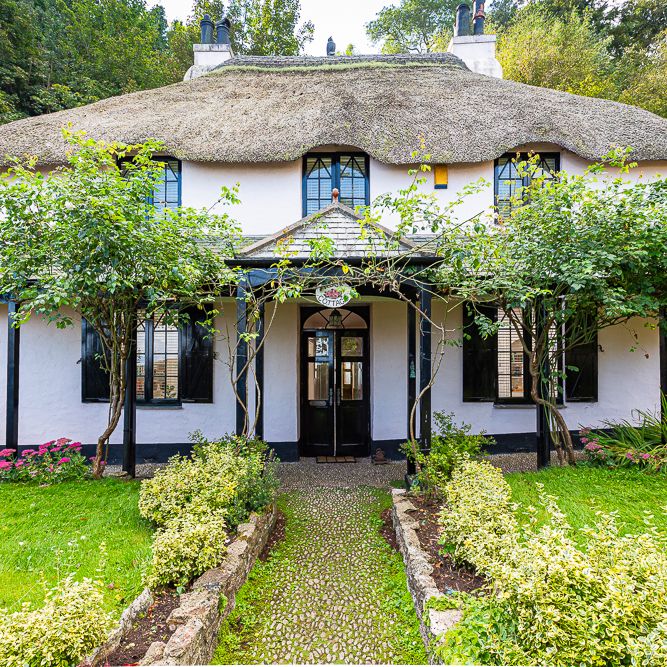 Charming white cottage with a thatched roof and black window frames, surrounded by lush greenery and a well-maintained garden path leading to the front door.