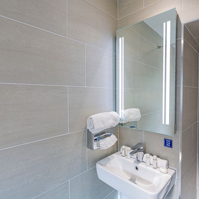 Modern bathroom with beige tiled walls, a rectangular sink, LED-lit mirror, towel rack with a rolled towel, and toiletry amenities on the sink.