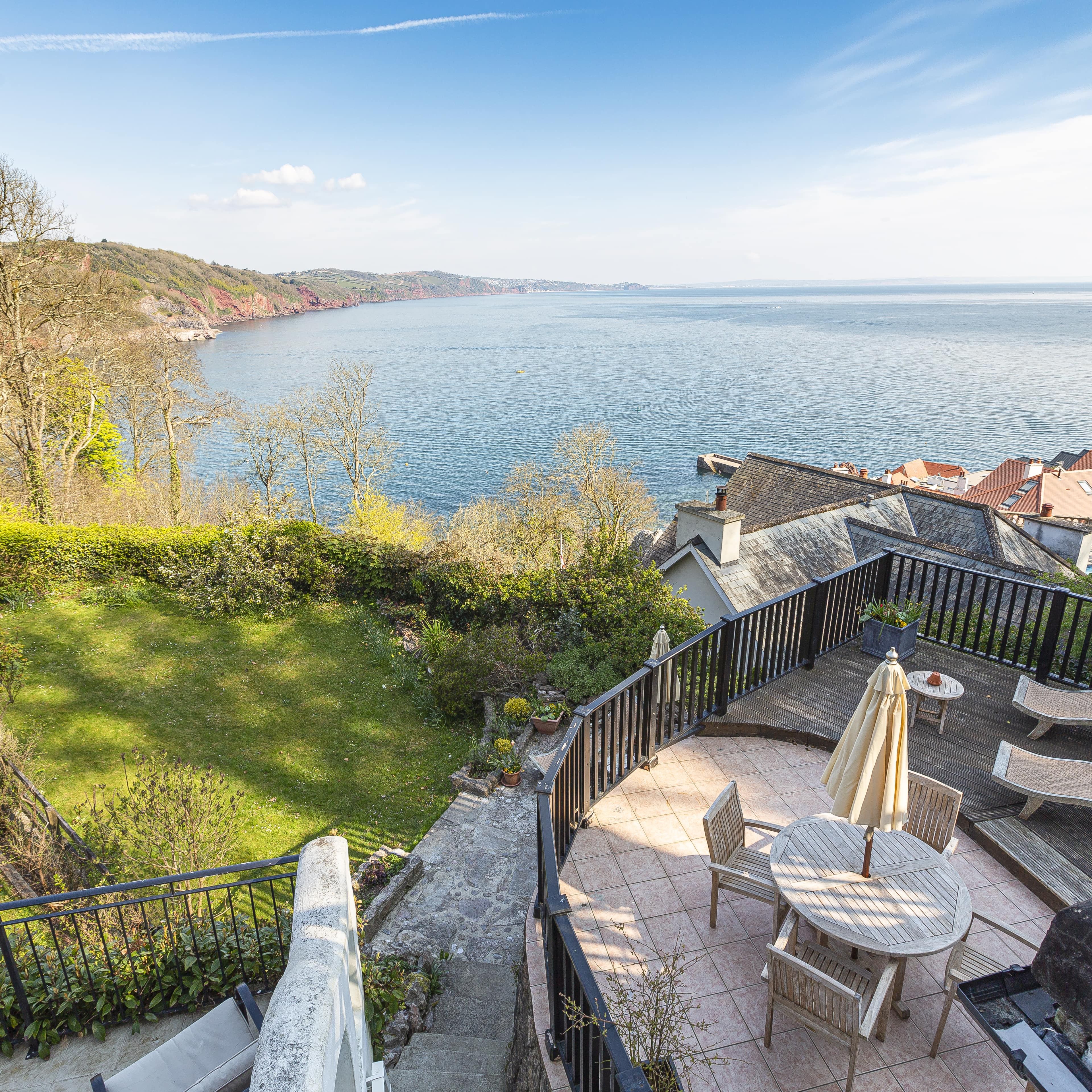 Scenic coastal view from an elevated terrace with outdoor seating, overlooking a garden, rooftops, and the ocean with a distant ship and coastline.