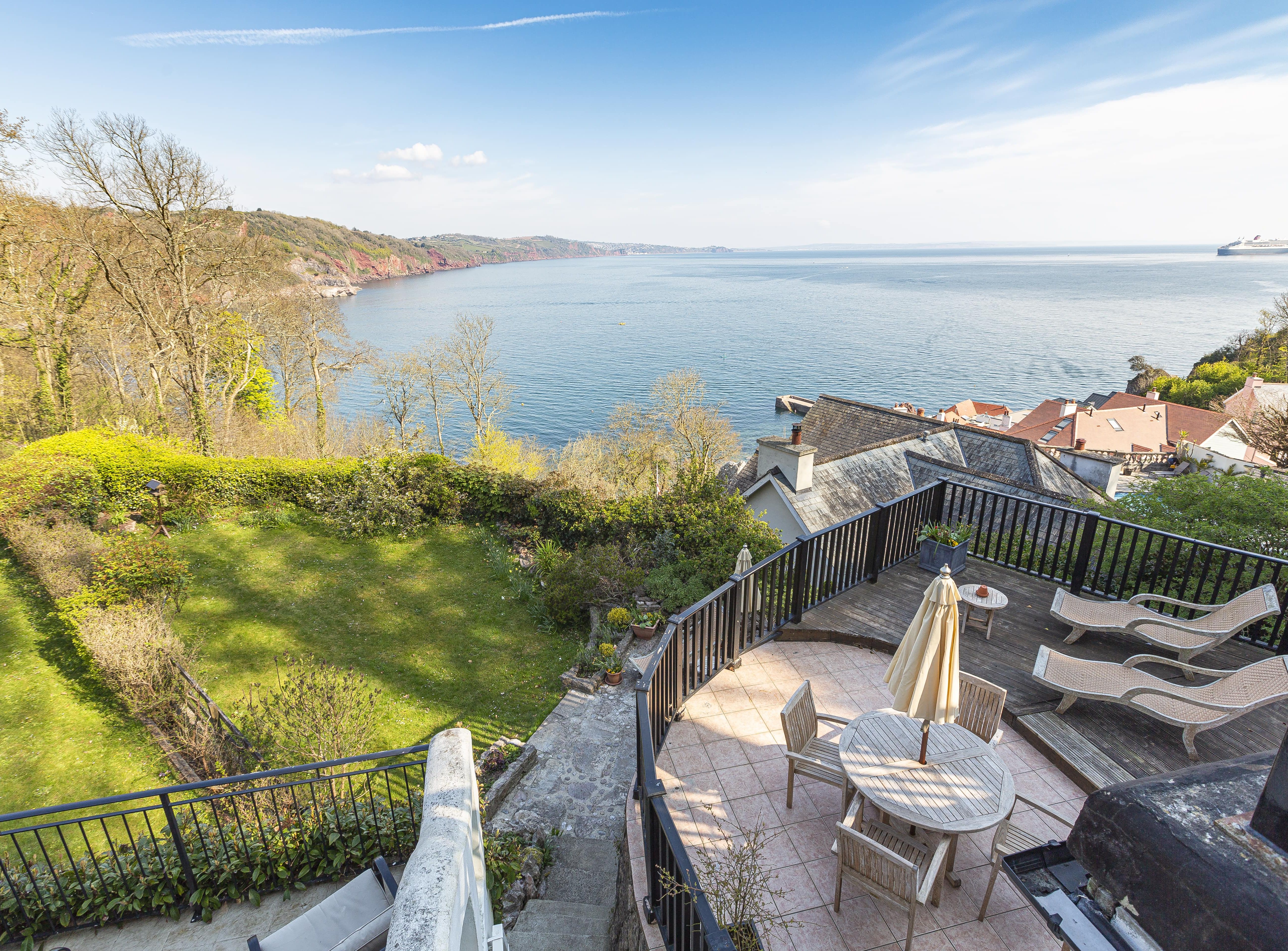 Scenic coastal view from an elevated terrace with outdoor seating, overlooking a garden, rooftops, and the ocean with a distant ship and coastline.