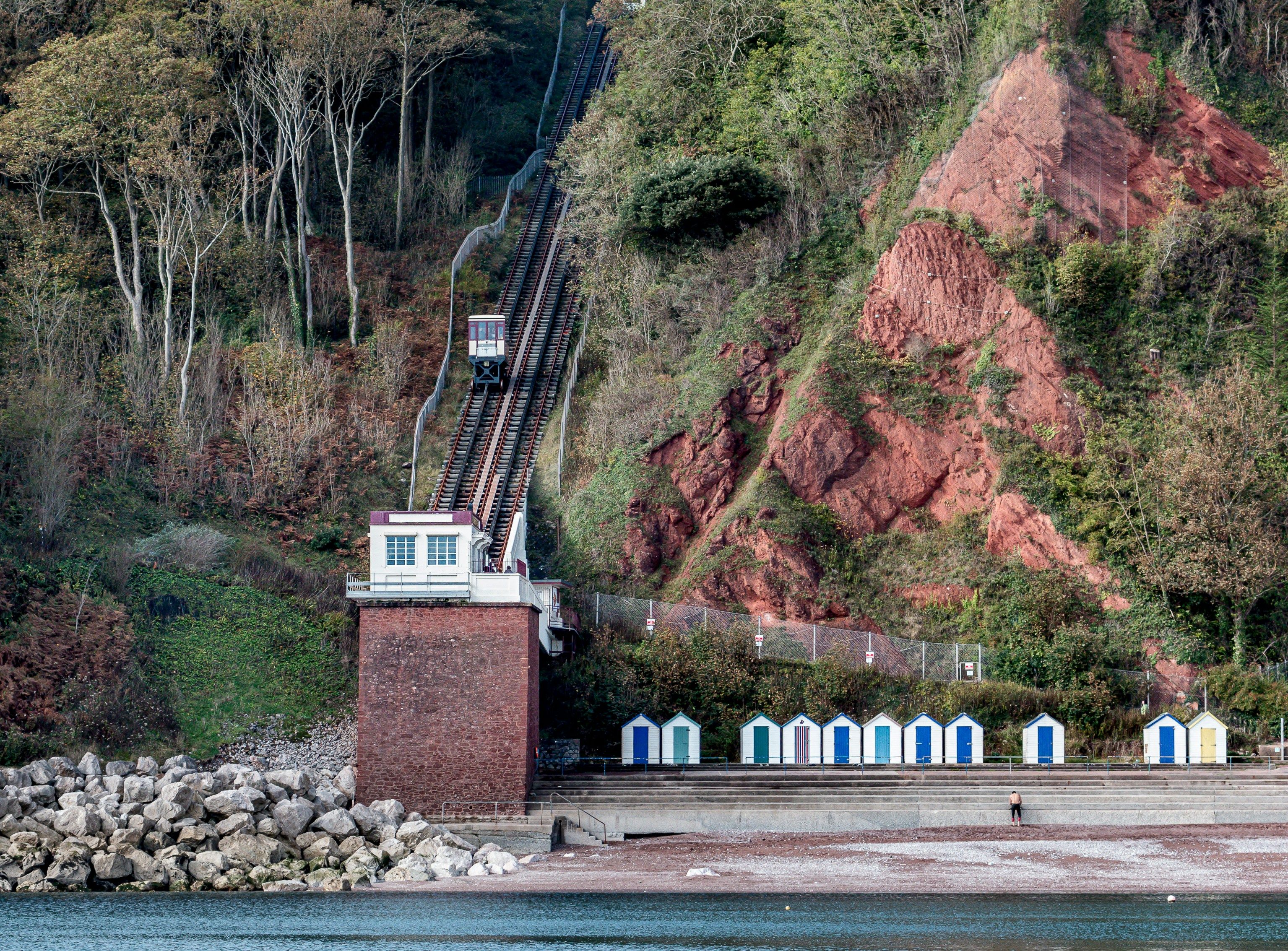 A funicular railway on a steep wooded hillside above beach huts and the shore.