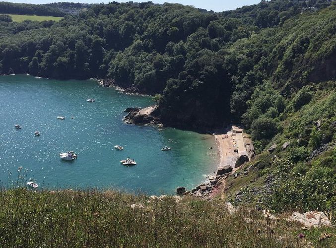 A small secluded cove with turquoise water, boats, and a sandy beach surrounded by steep green hills.