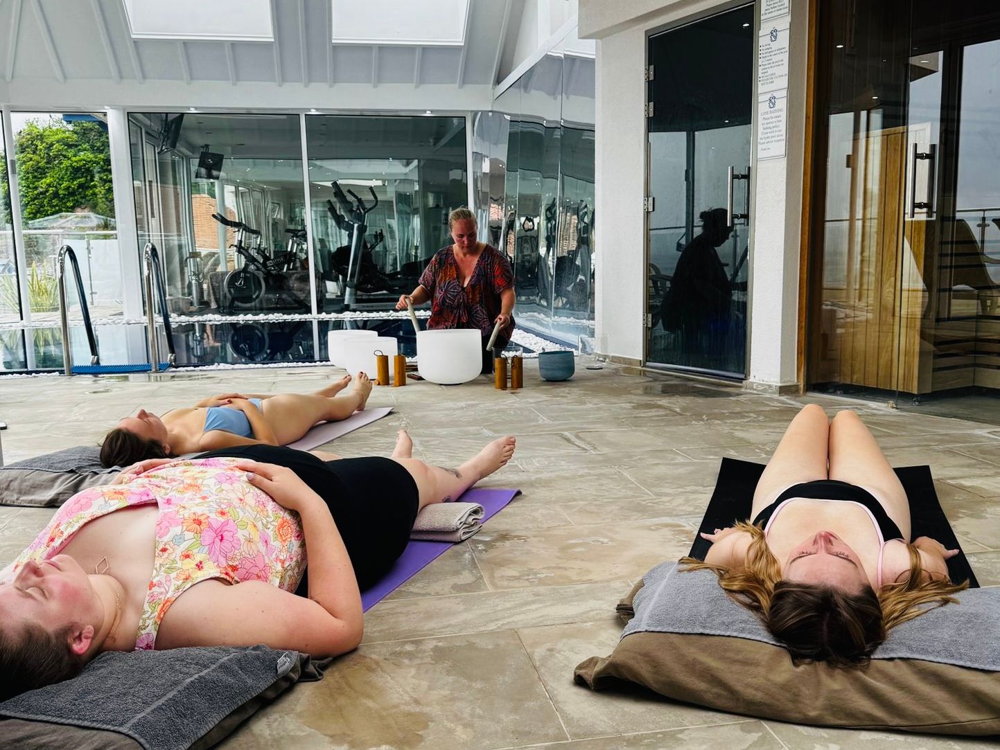 Group of women lying on yoga mats indoors during a sound bath session led by an instructor with crystal bowls.