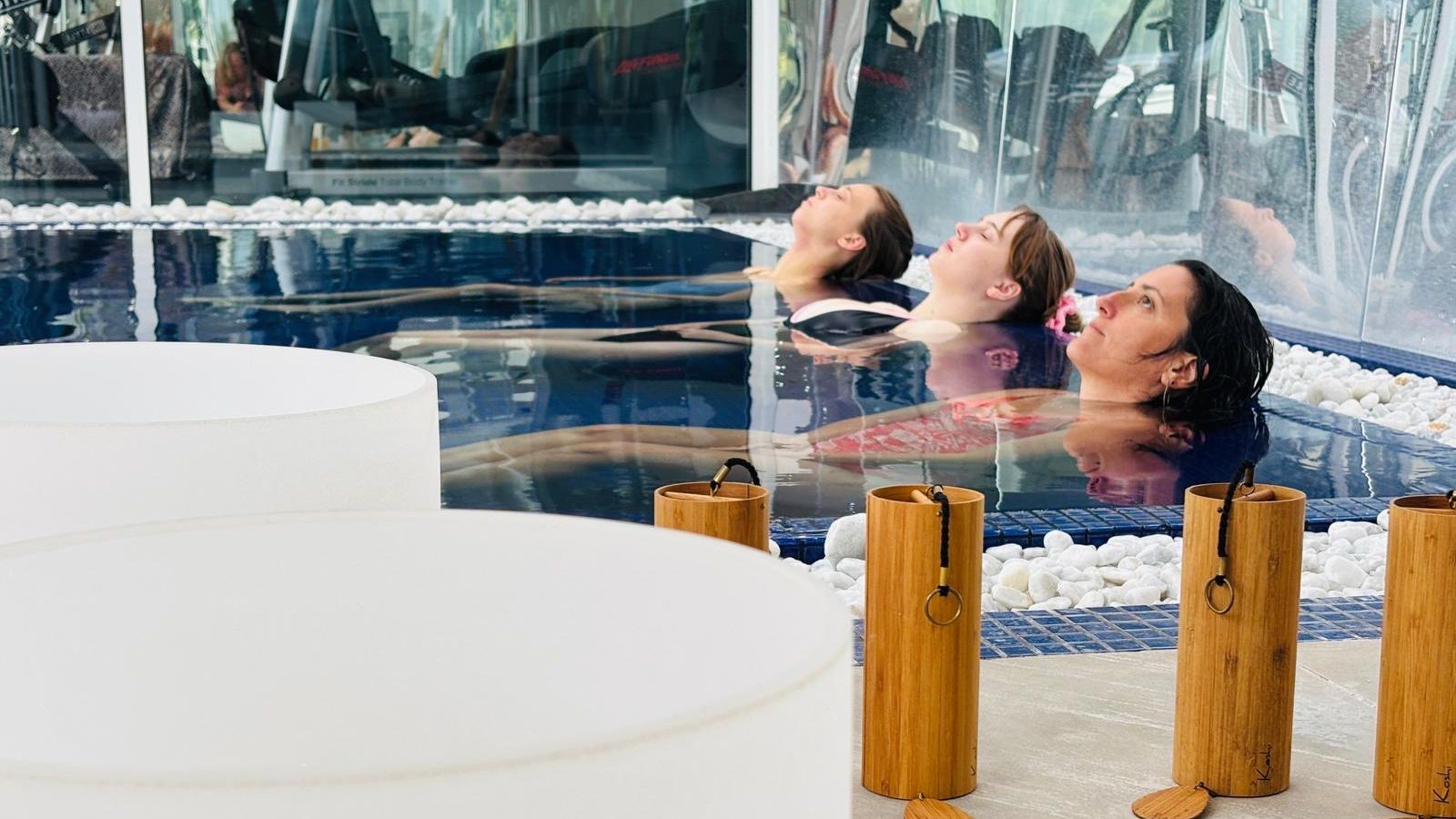 Three women relaxing and floating in a spa pool next to bamboo and crystal singing bowls.