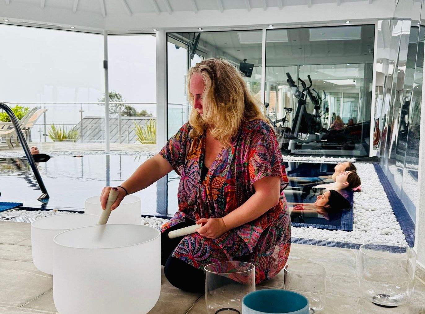 Woman playing crystal singing bowls with people relaxing in a pool nearby in a bright indoor space.
