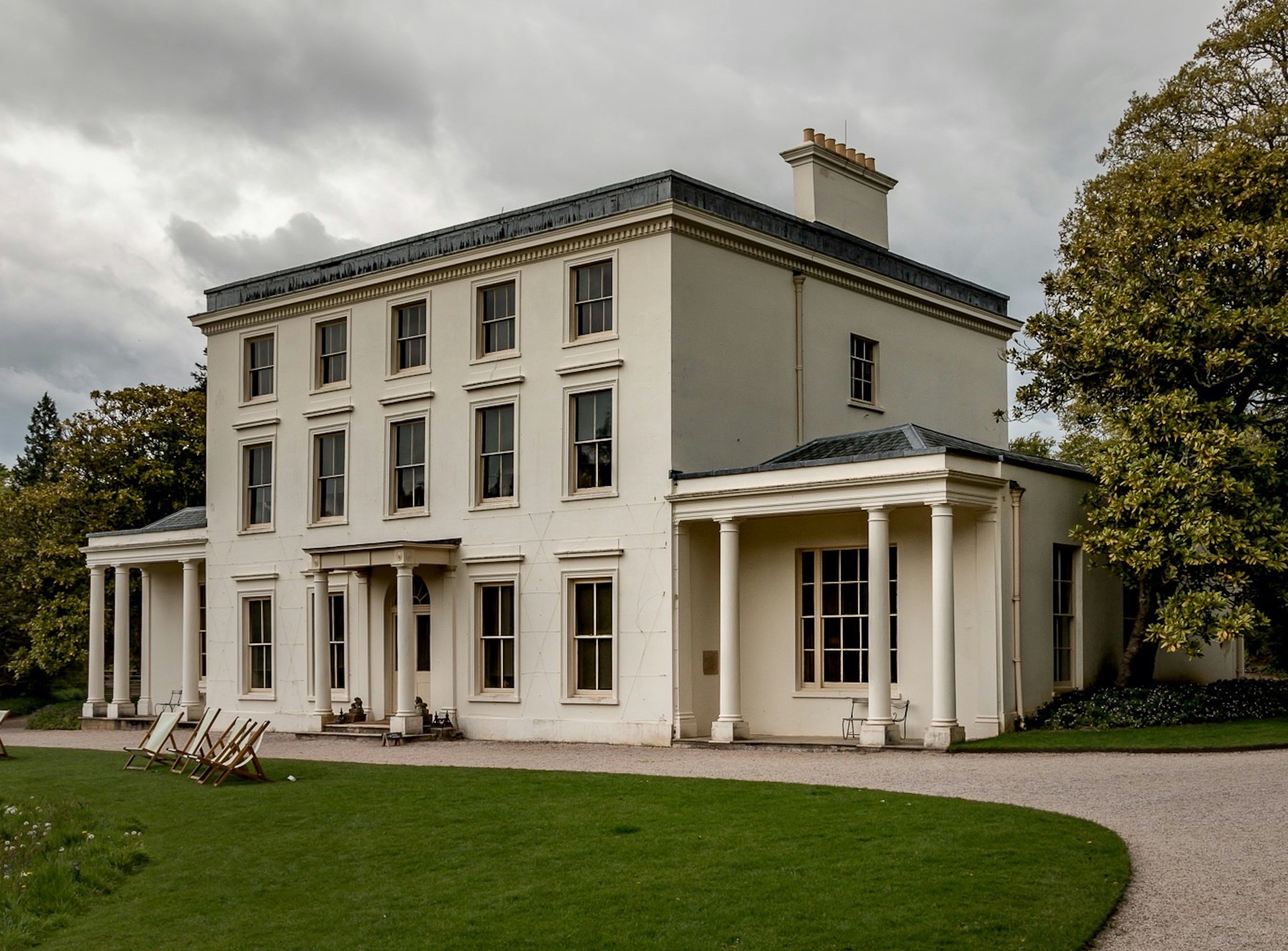 Large white Georgian-style house with columns and a gravel driveway surrounded by trees and green lawn
