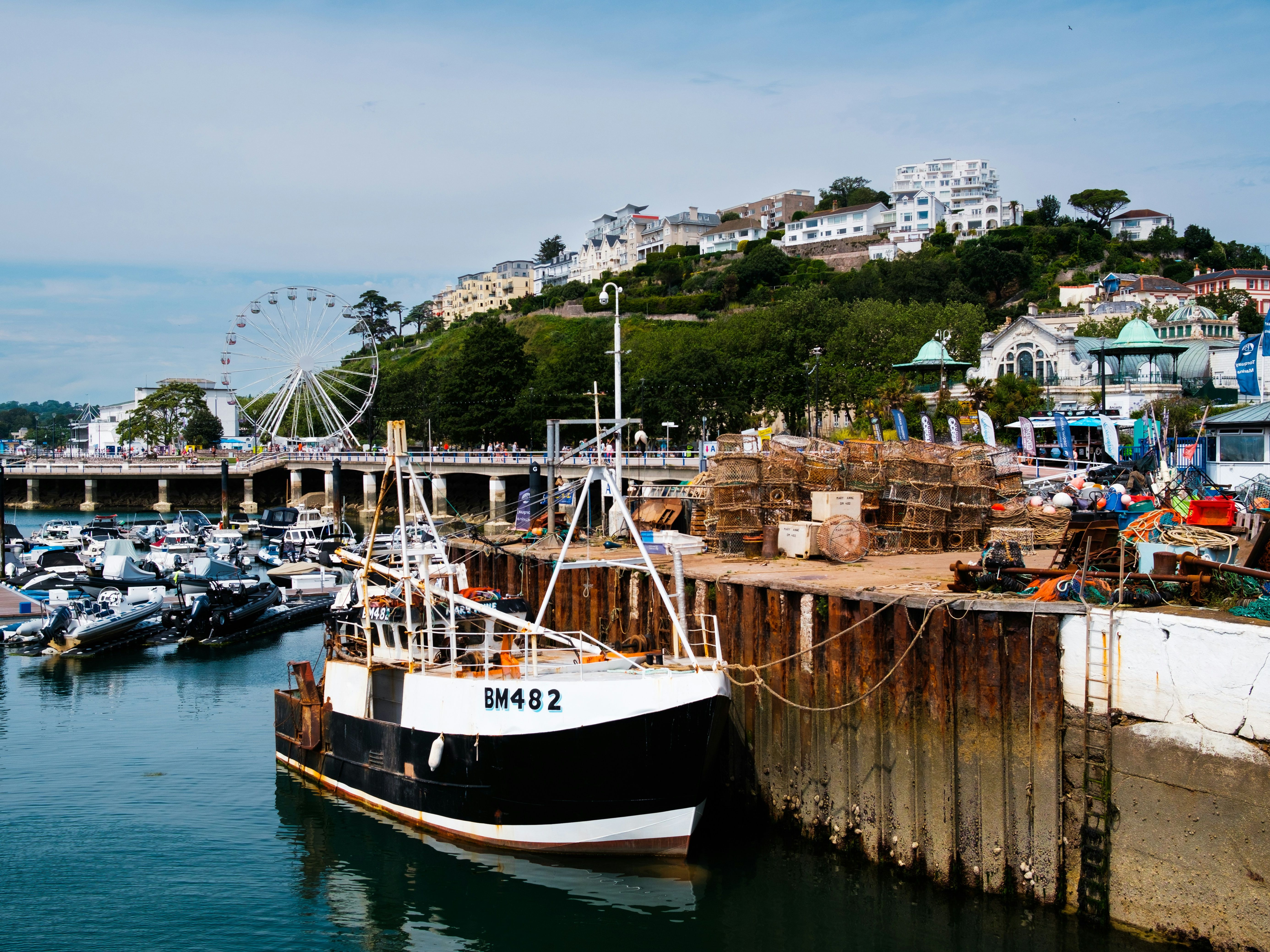 Fishing boat docked at a marina with lobster pots and fishing gear stacked on the quay, a ferris wheel, and hillside buildings in the background.