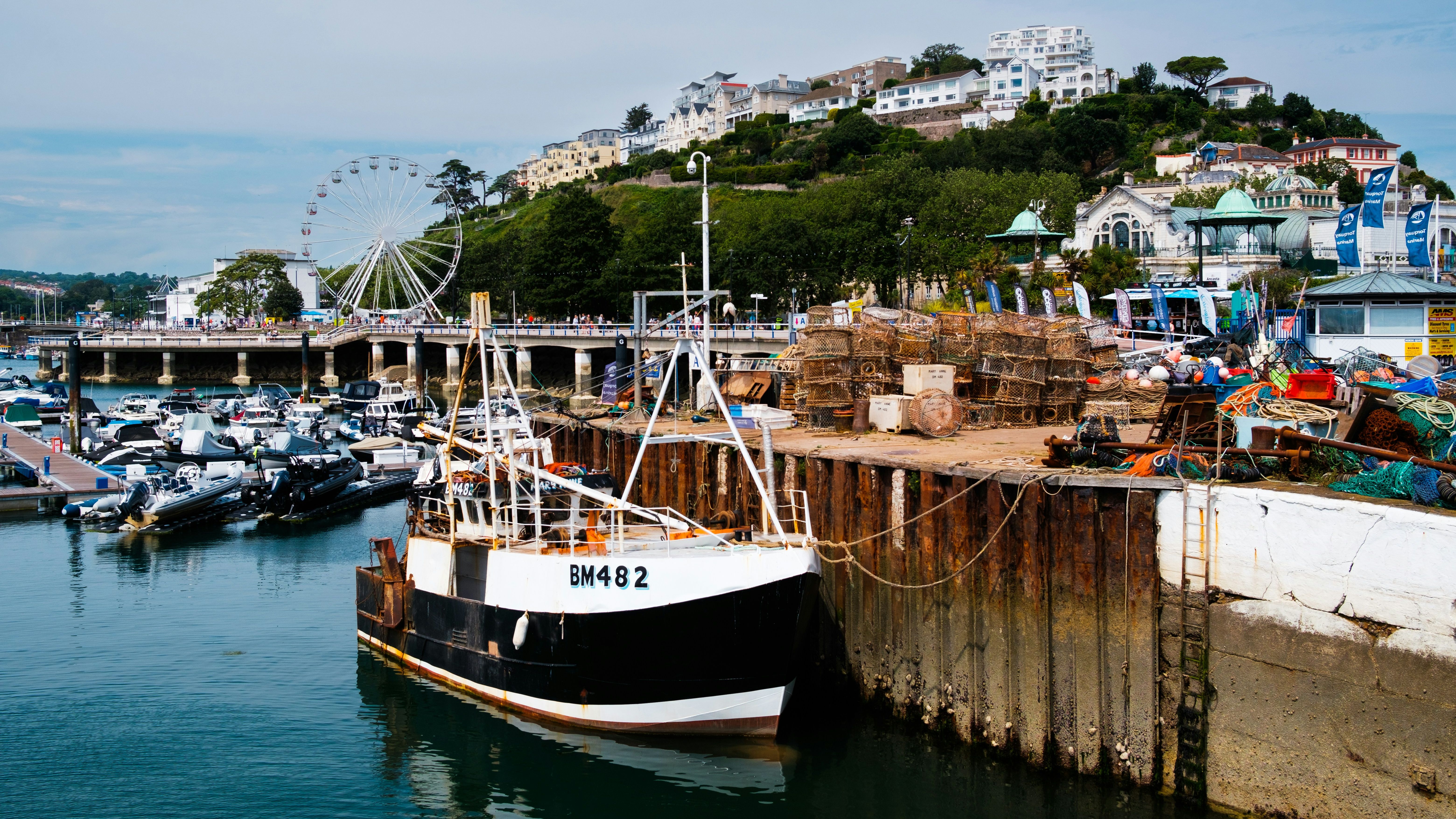 Fishing boat docked at a marina with lobster pots and fishing gear stacked on the quay, a ferris wheel, and hillside buildings in the background.
