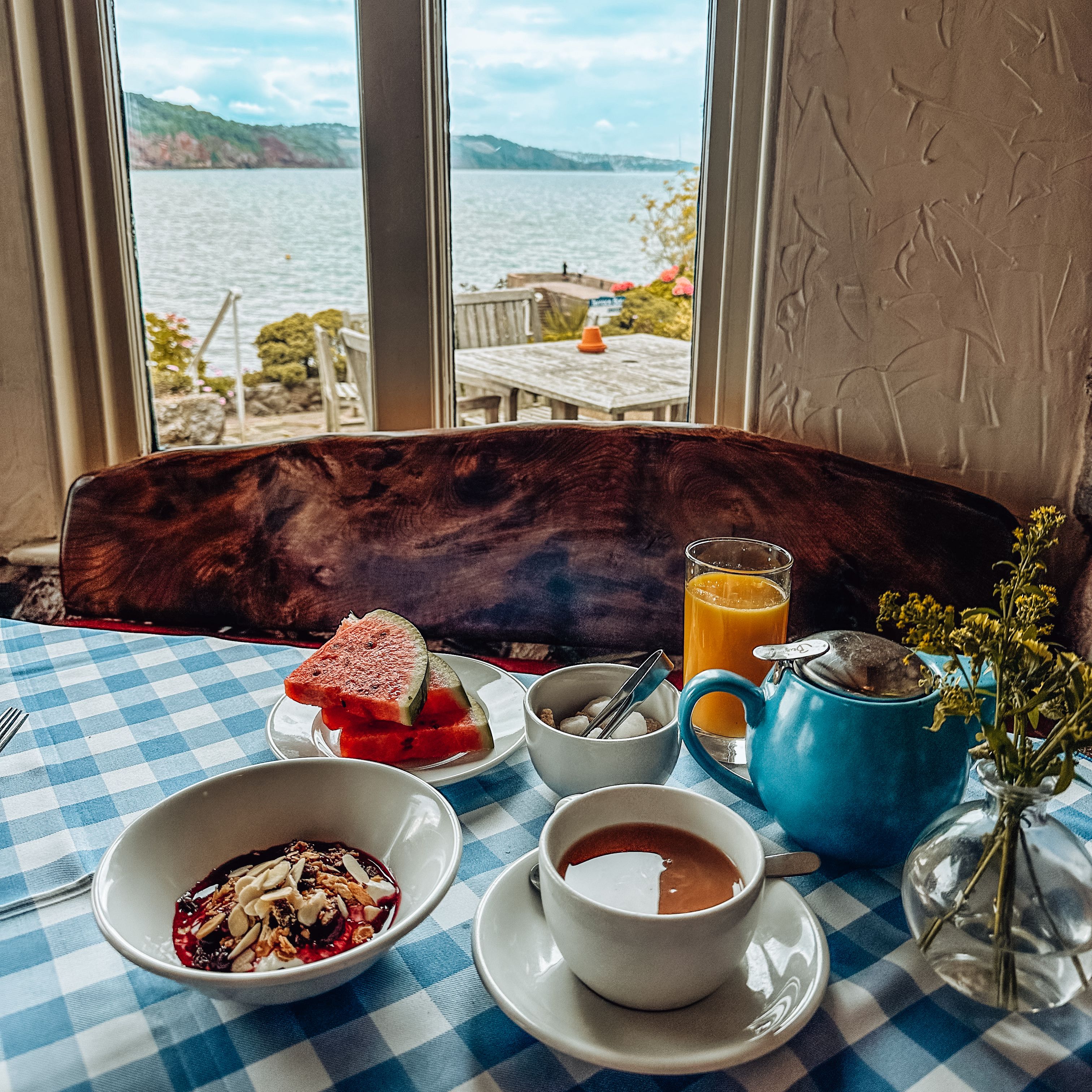 Breakfast table with tea, juice, fruit, and yogurt by a window overlooking a lake.
