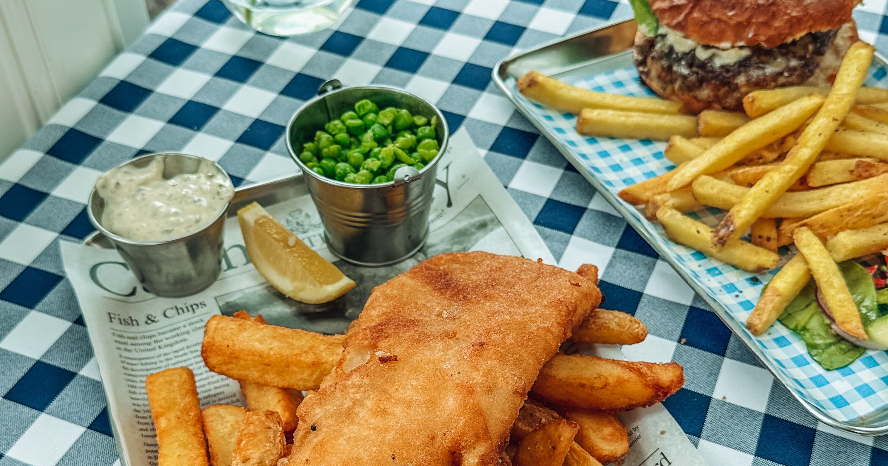 A tray of fish and chips with peas, tartar sauce, and a lemon wedge on a checkered tablecloth, with a burger and fries in the background.