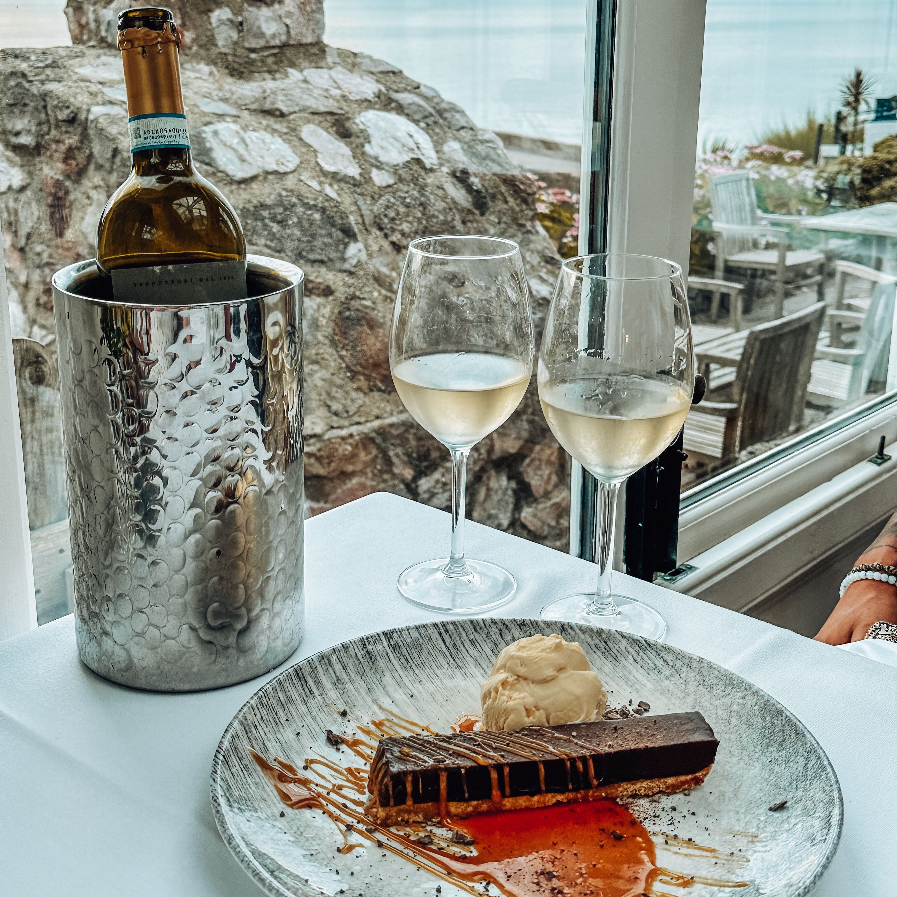 A table set with a dessert plate, two glasses of white wine, and a wine bottle in a chiller, next to a window with a scenic view of water and outdoor seating.