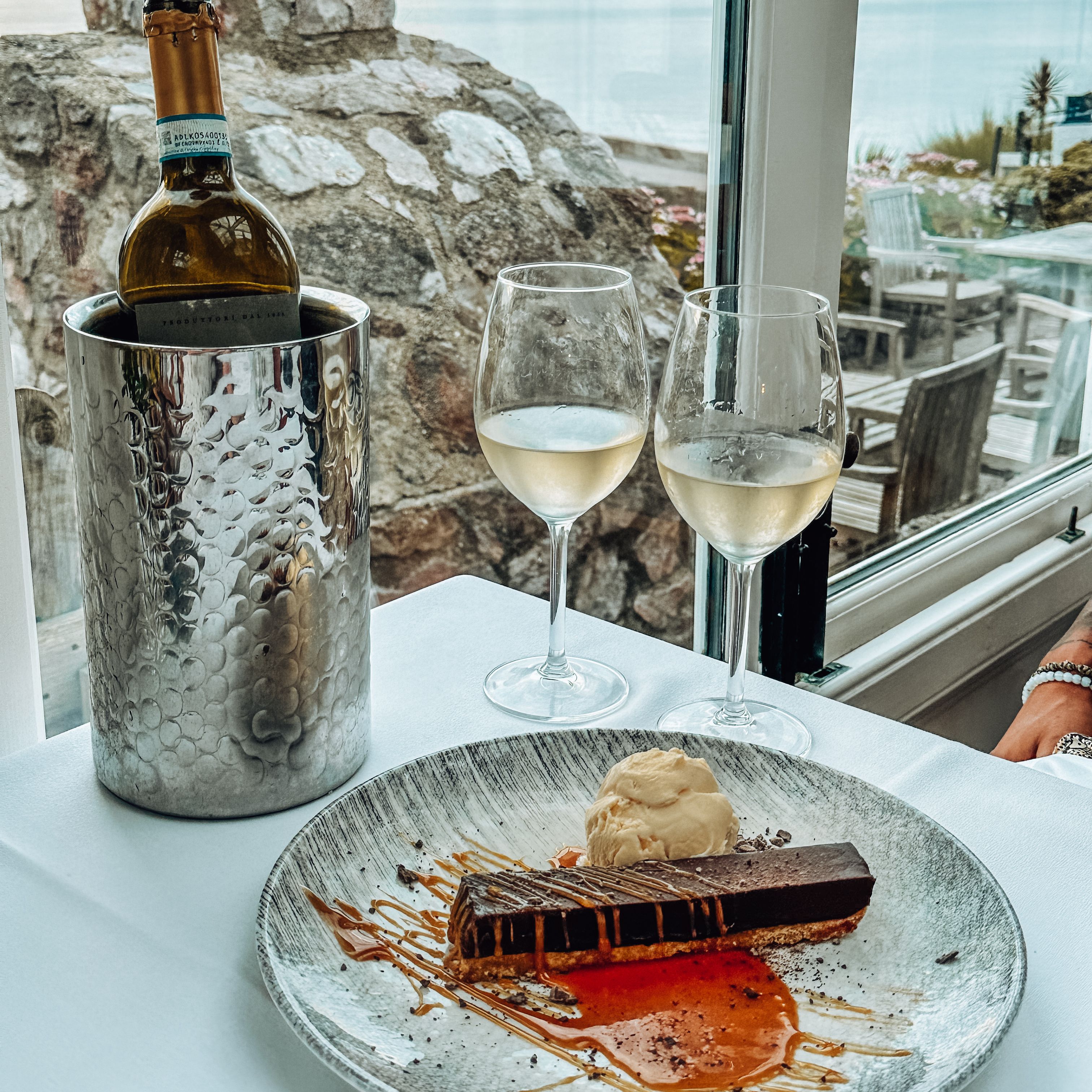 A table set with a dessert plate, two glasses of white wine, and a wine bottle in a chiller, next to a window with a scenic view of water and outdoor seating.