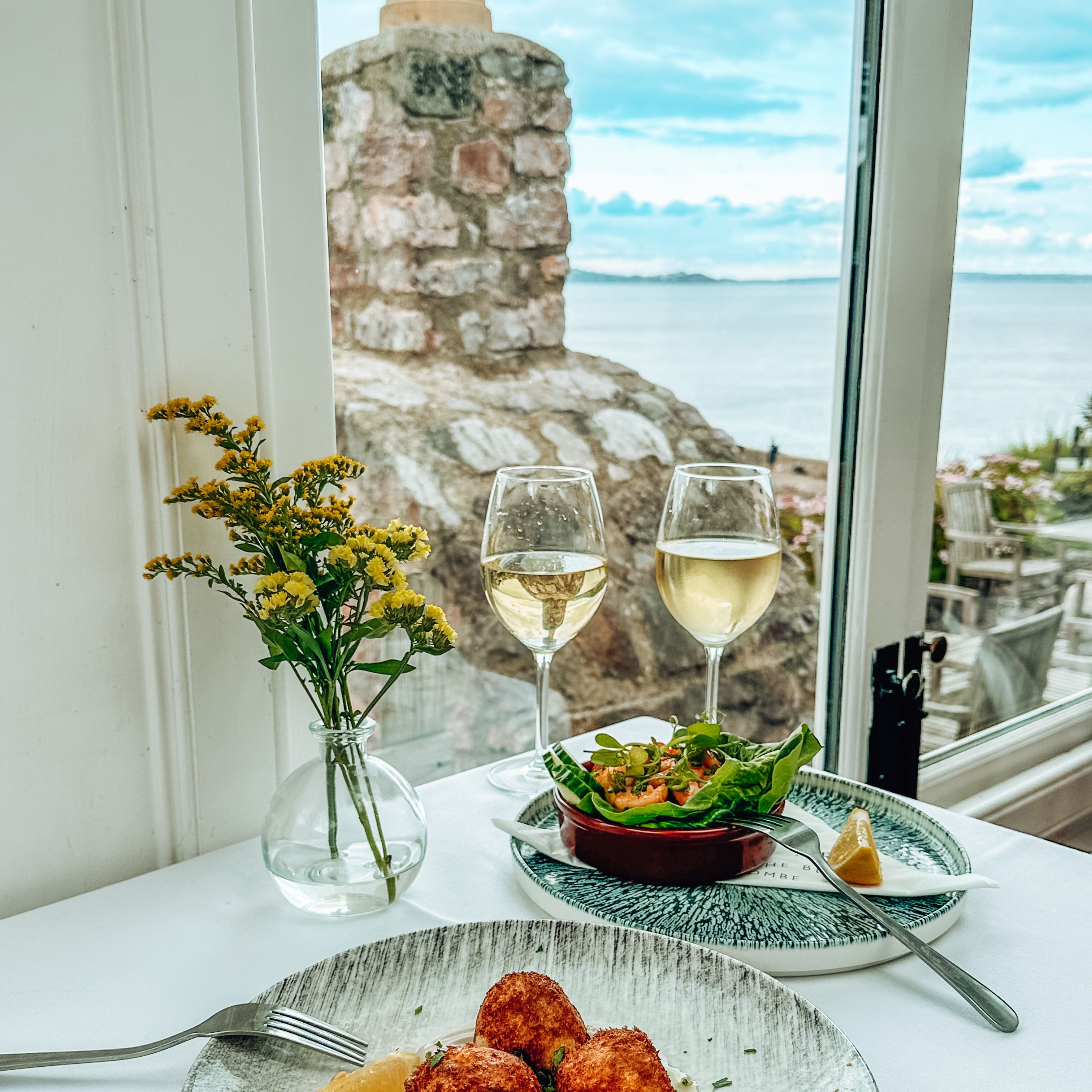 Table setting with seafood appetizer, white wine, salad, and a view of the sea through a window.