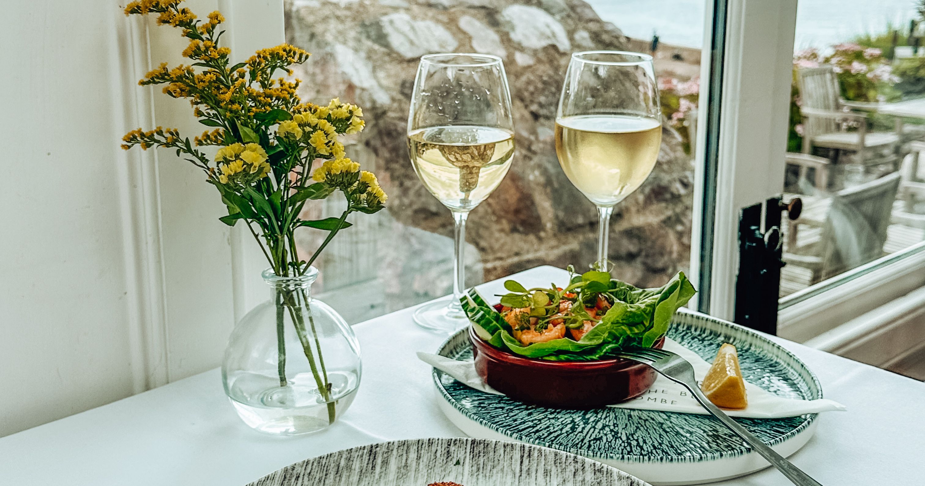 Table setting with seafood appetizer, white wine, salad, and a view of the sea through a window.