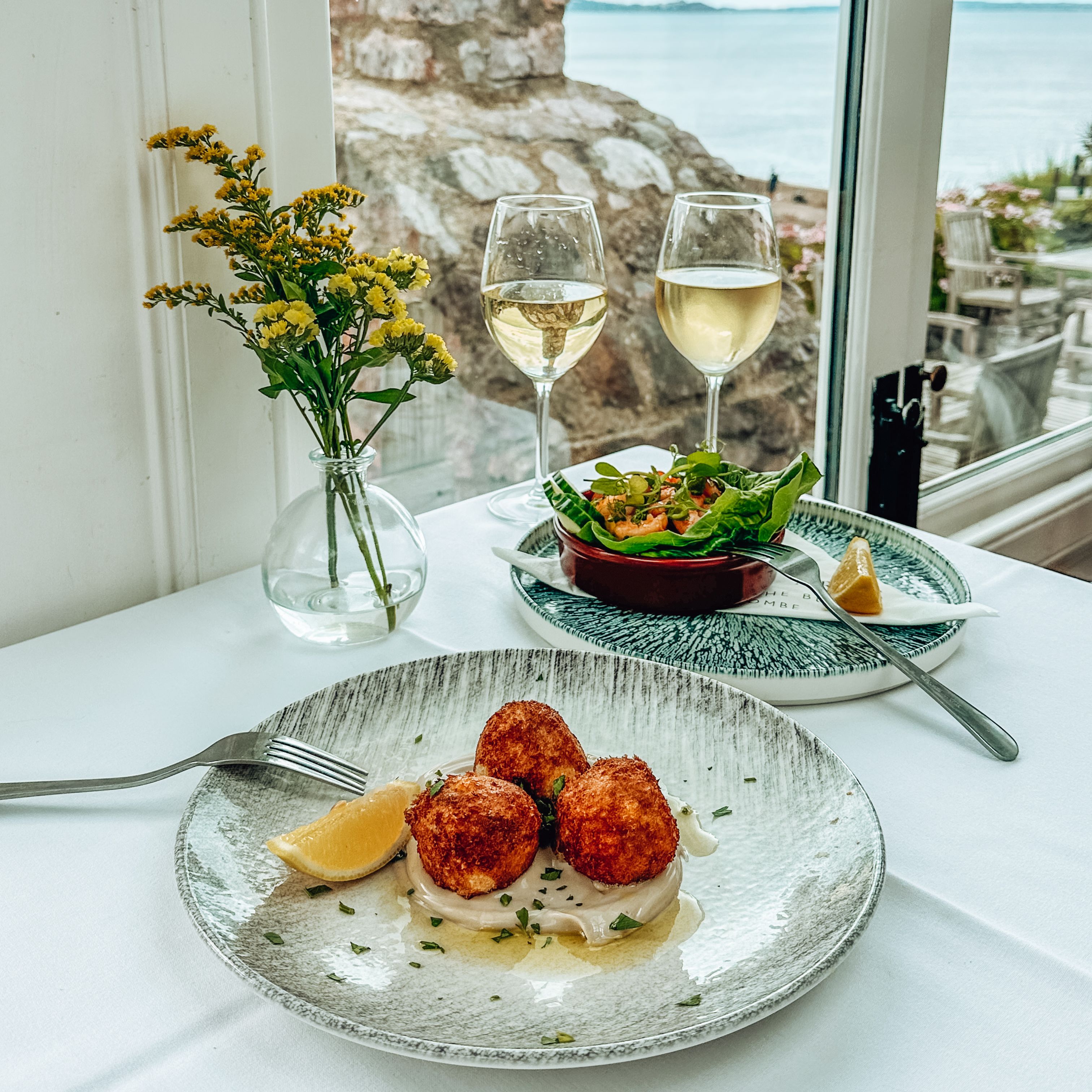 Table setting with seafood appetizer, white wine, salad, and a view of the sea through a window.
