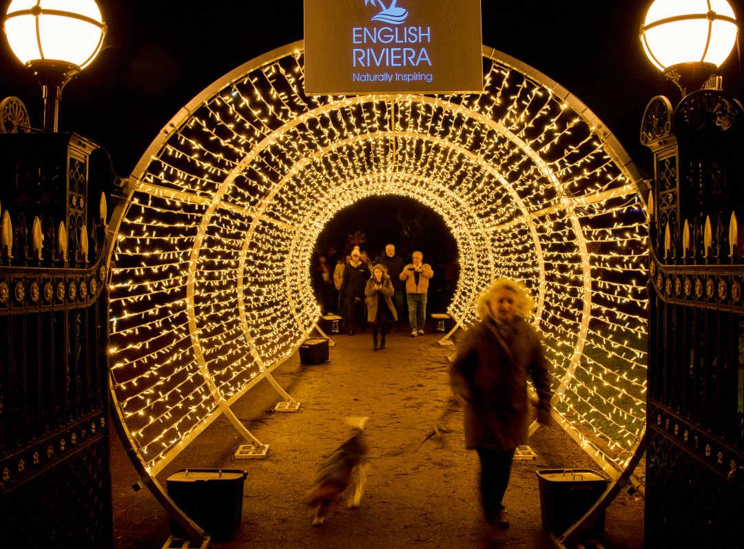 People walking through a tunnel decorated with golden fairy lights at night, under a sign that reads 'English Riviera'.