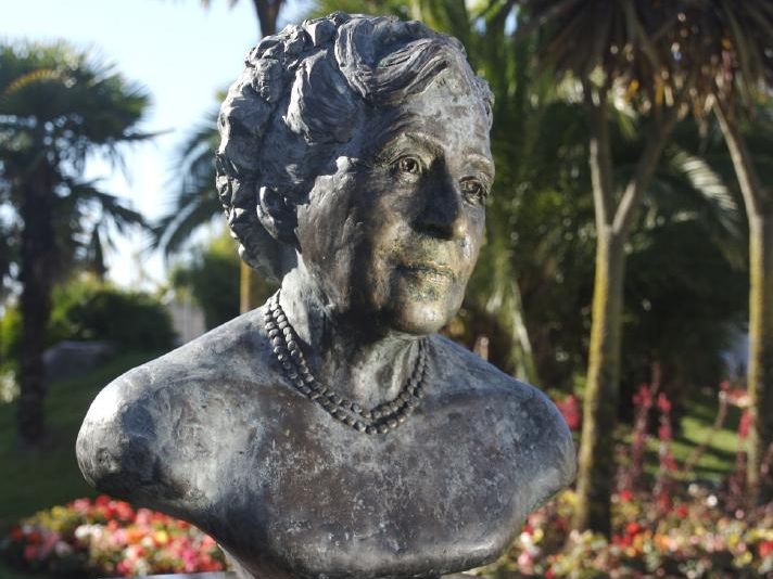 Bust statue of Agatha Christie on a pedestal with flowers and palm trees in the background.