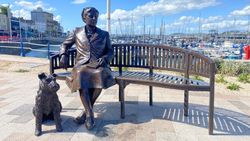 Bronze statue of a woman sitting on a bench with a dog at her feet, located by a marina with boats and buildings in the background.