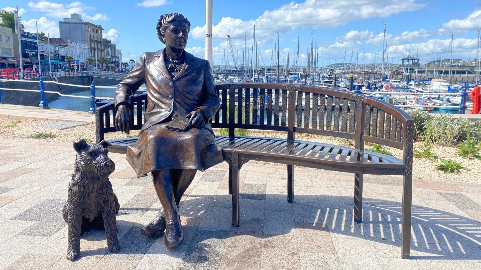 Bronze statue of a woman sitting on a bench with a dog at her feet, located by a marina with boats and buildings in the background.