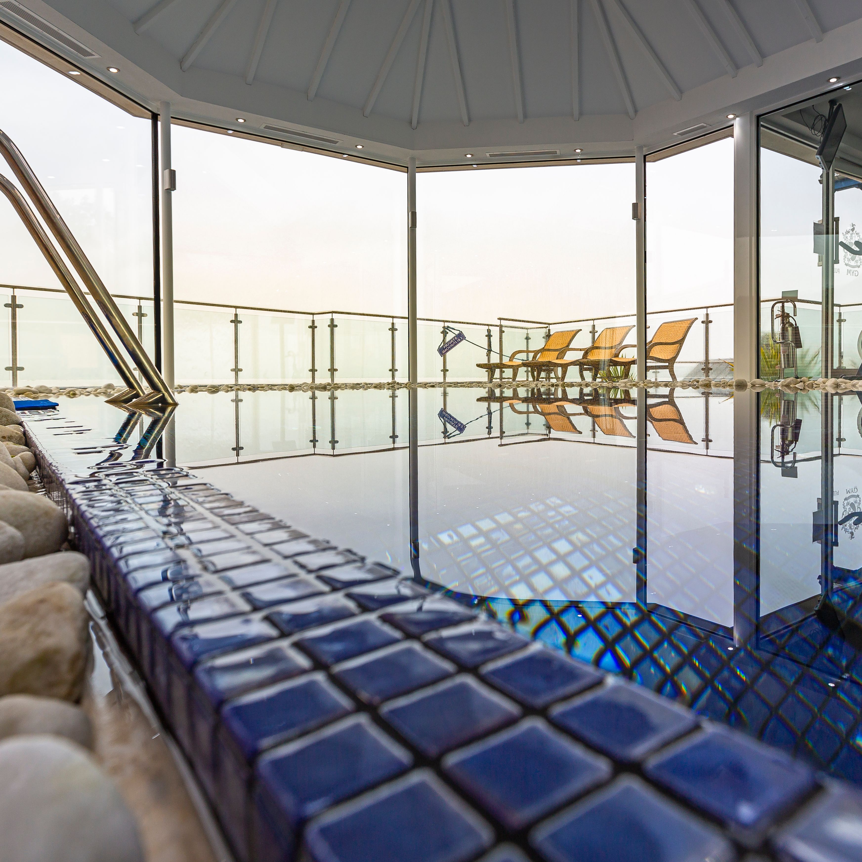 Indoor spa pool area with blue tiles, exercise equipment, and lounge chairs by large glass windows.