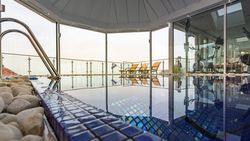 Indoor spa pool area with blue tiles, exercise equipment, and lounge chairs by large glass windows.