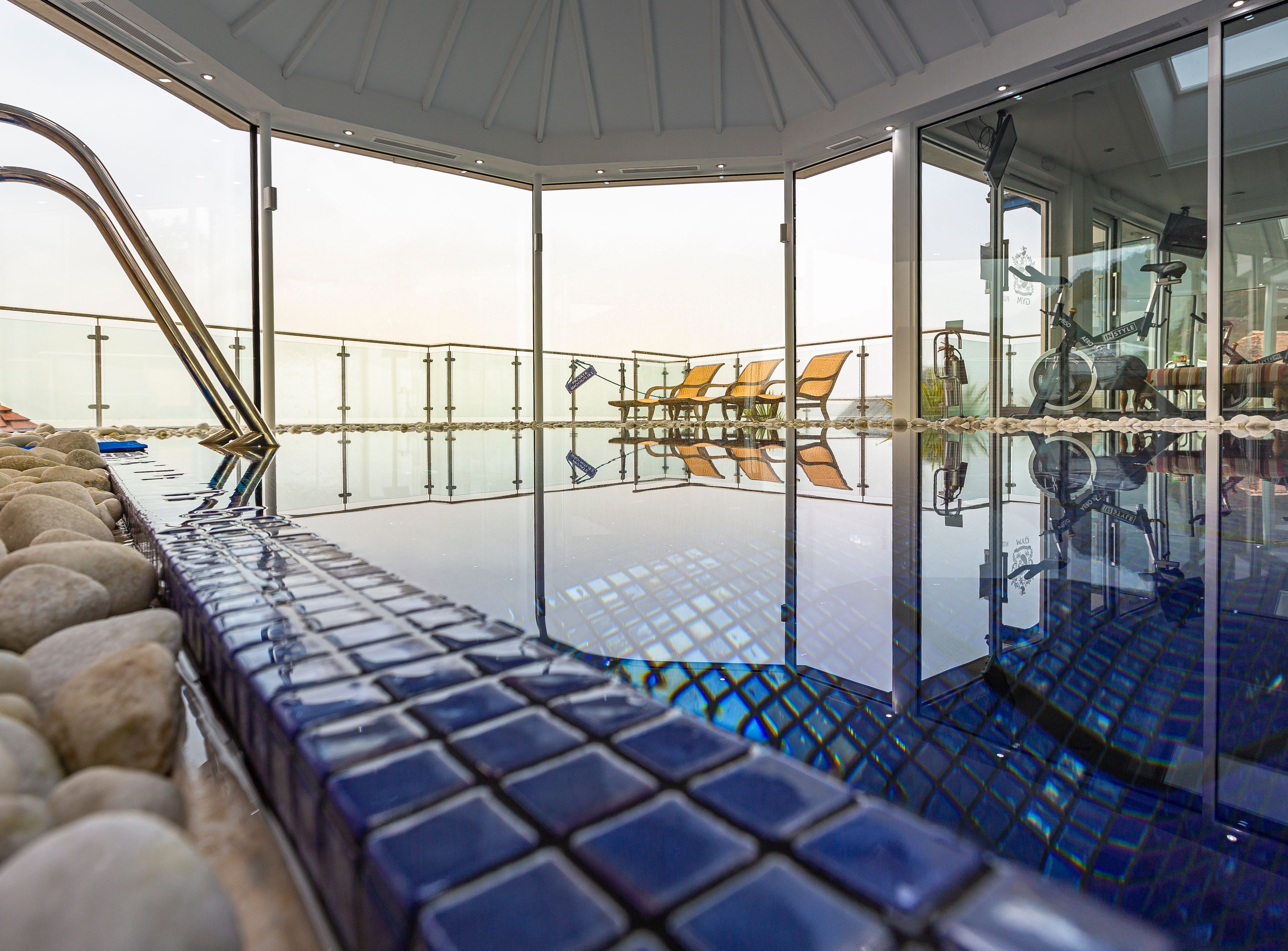 Indoor spa pool area with blue tiles, exercise equipment, and lounge chairs by large glass windows.