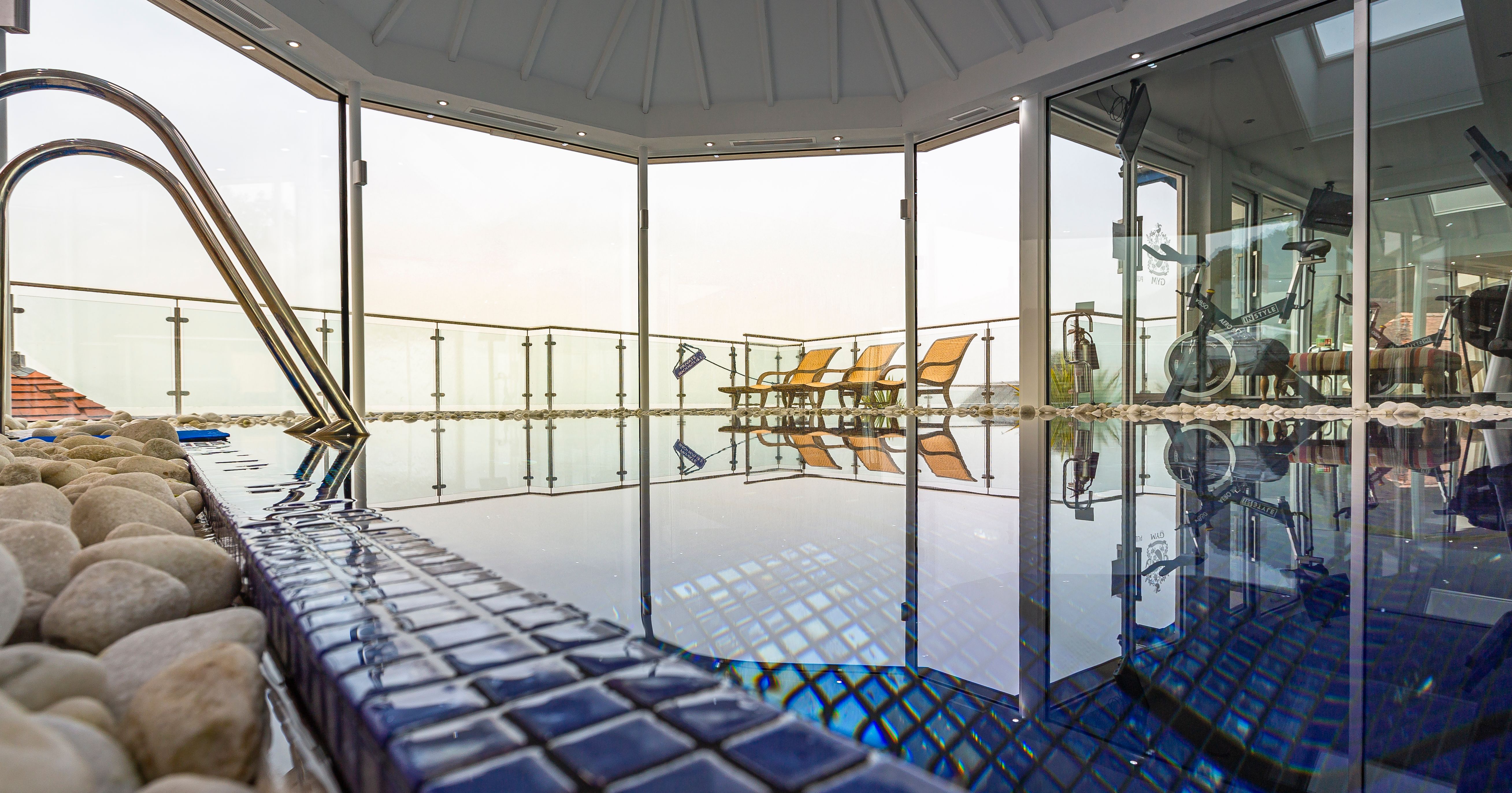 Indoor spa pool area with blue tiles, exercise equipment, and lounge chairs by large glass windows.