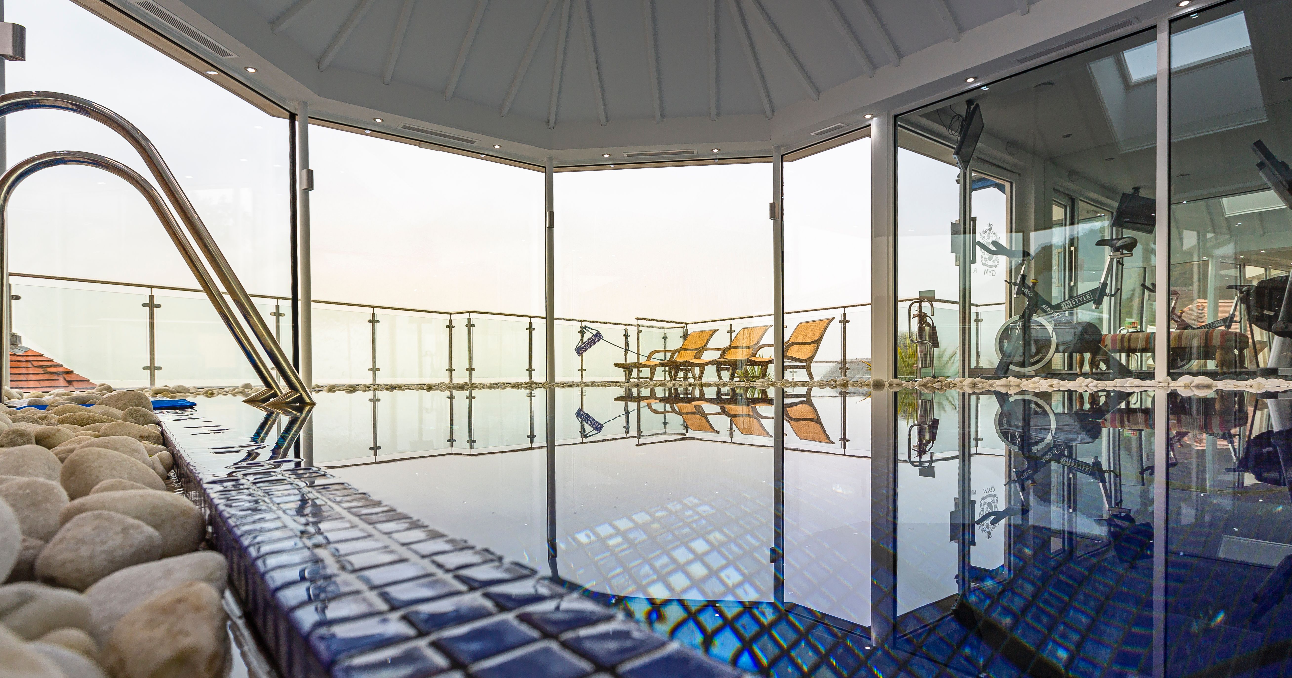 Indoor spa pool area with blue tiles, exercise equipment, and lounge chairs by large glass windows.