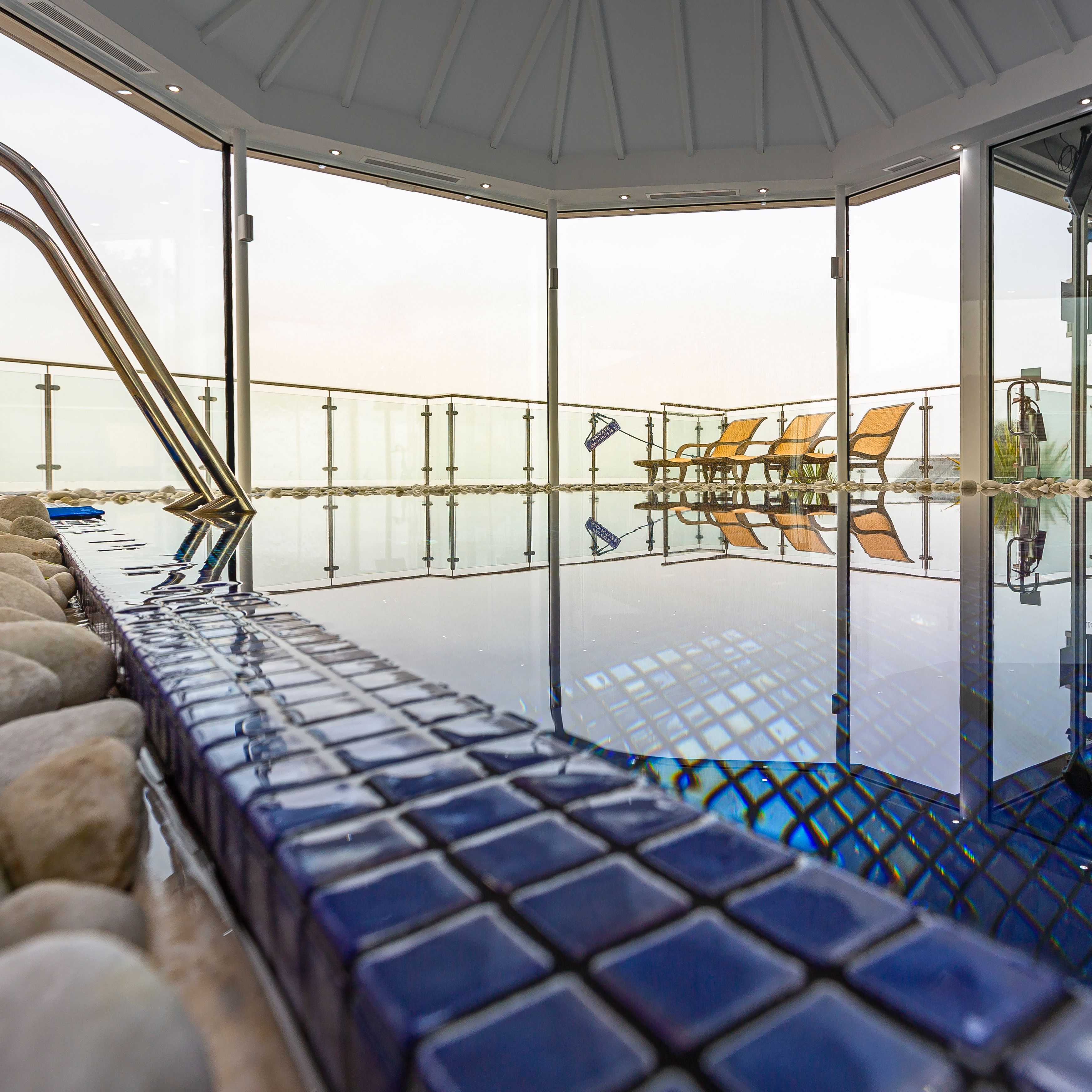 Indoor spa pool area with blue tiles, exercise equipment, and lounge chairs by large glass windows.