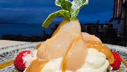 Poached pear dessert with caramel sauce, whipped cream, mint leaf garnish, and raspberries on a plate.