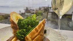A gourmet vegetarian dish with pastry, mashed potatoes, broccoli, and brown sauce, served alongside a glass of white wine with a waterfront view in the background.