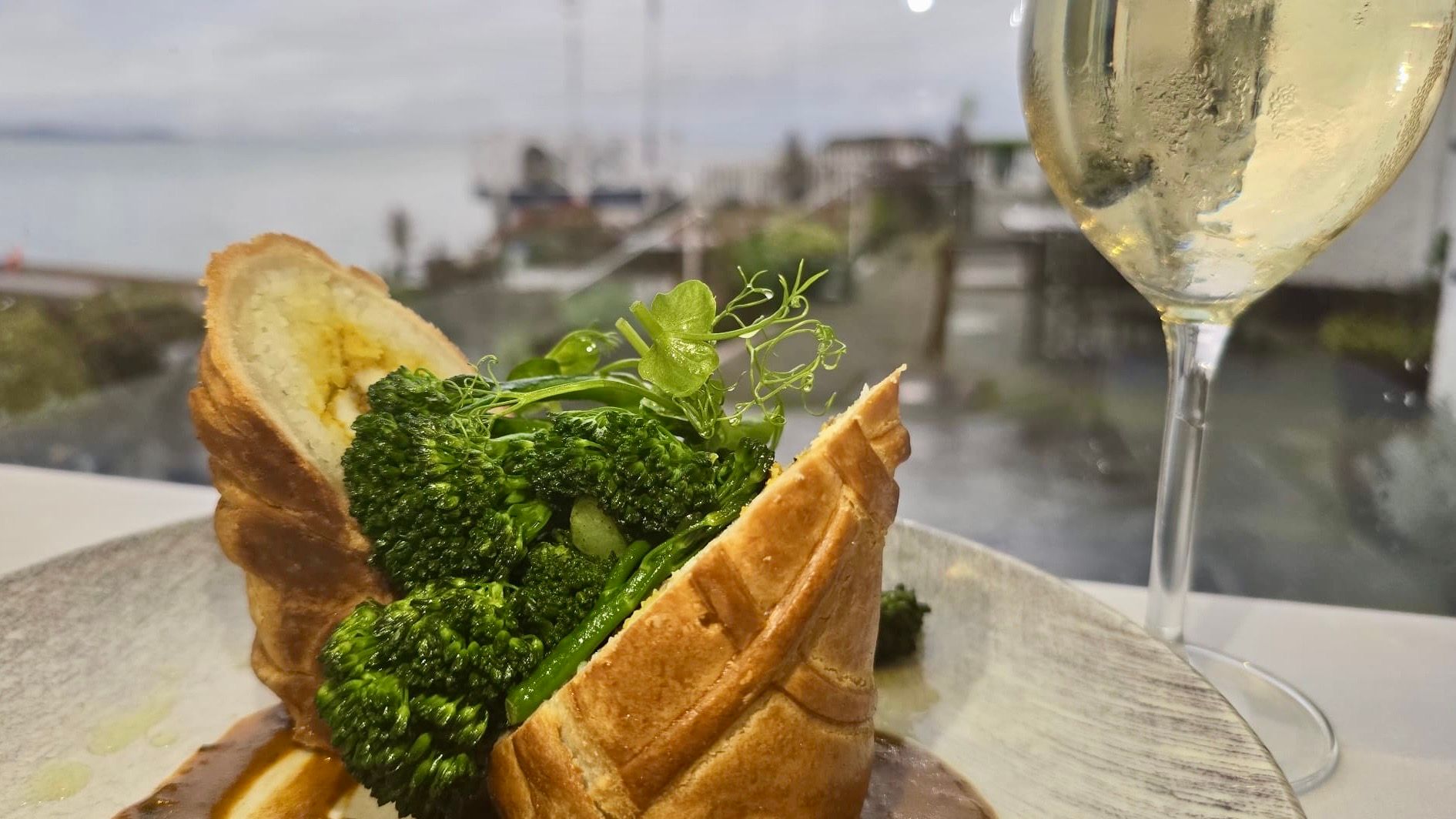 A gourmet vegetarian dish with pastry, mashed potatoes, broccoli, and brown sauce, served alongside a glass of white wine with a waterfront view in the background.