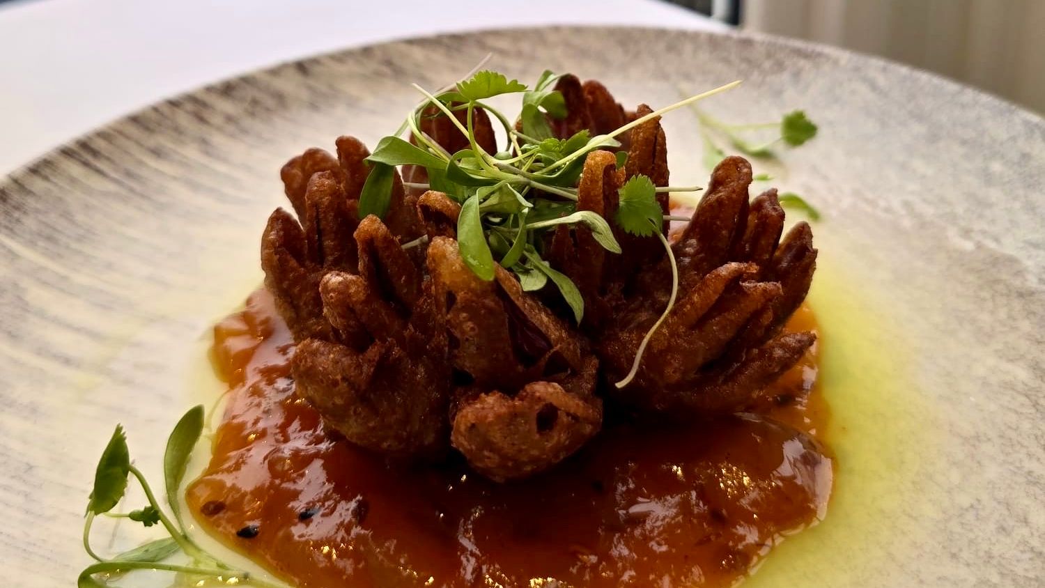 Plated gourmet dish resembling a blooming onion atop a sauce, garnished with microgreens, with a scenic seaside view in the background.