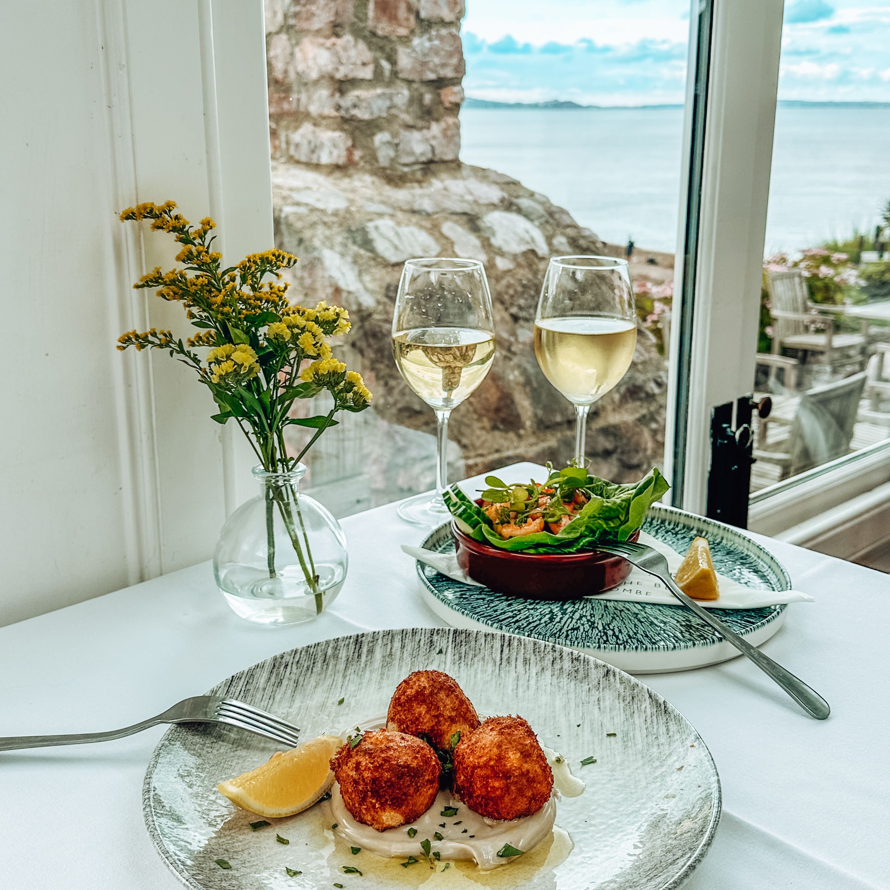 Table set with gourmet seafood appetizers, white wine glasses, and a seaside view.