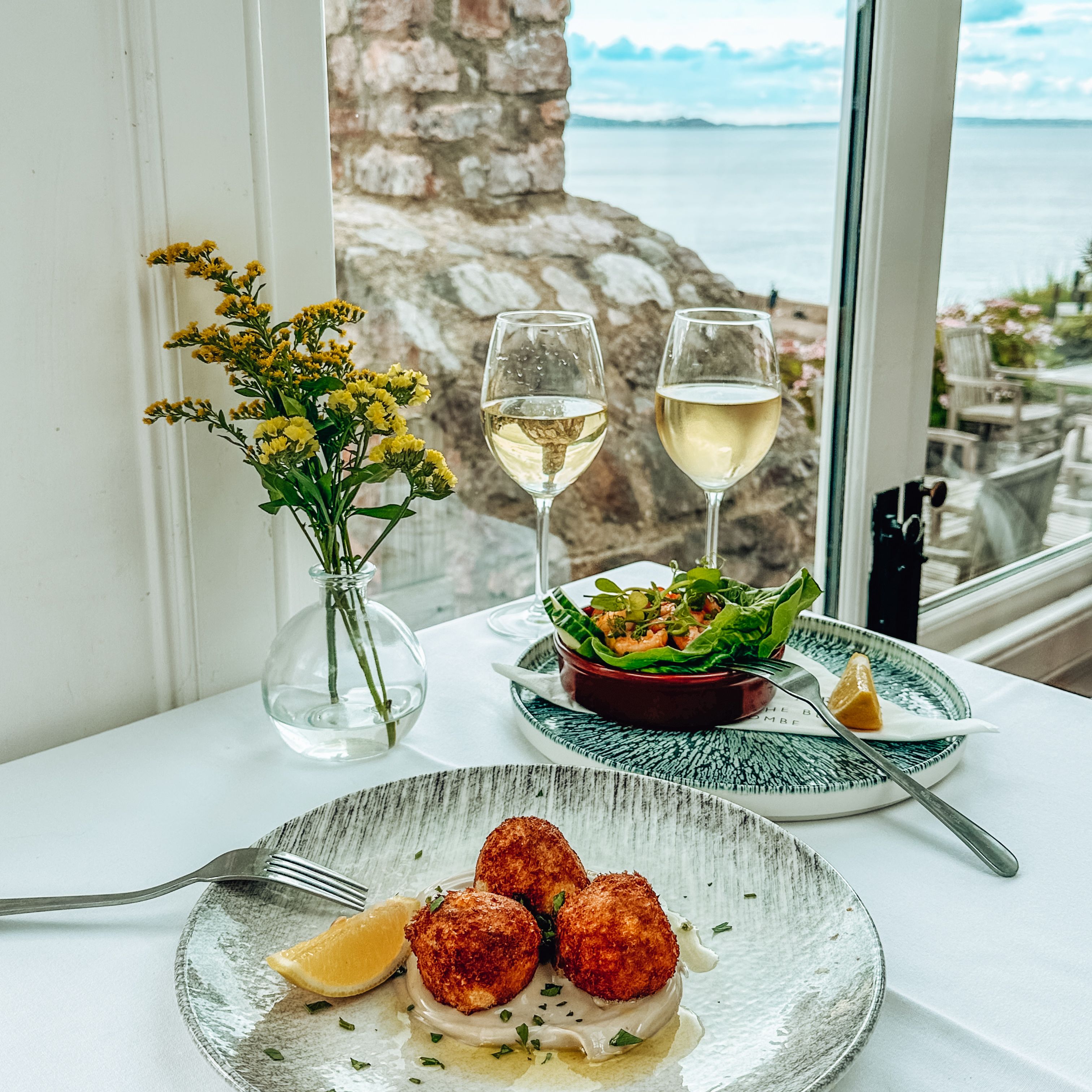Table set with gourmet seafood appetizers, white wine glasses, and a seaside view.