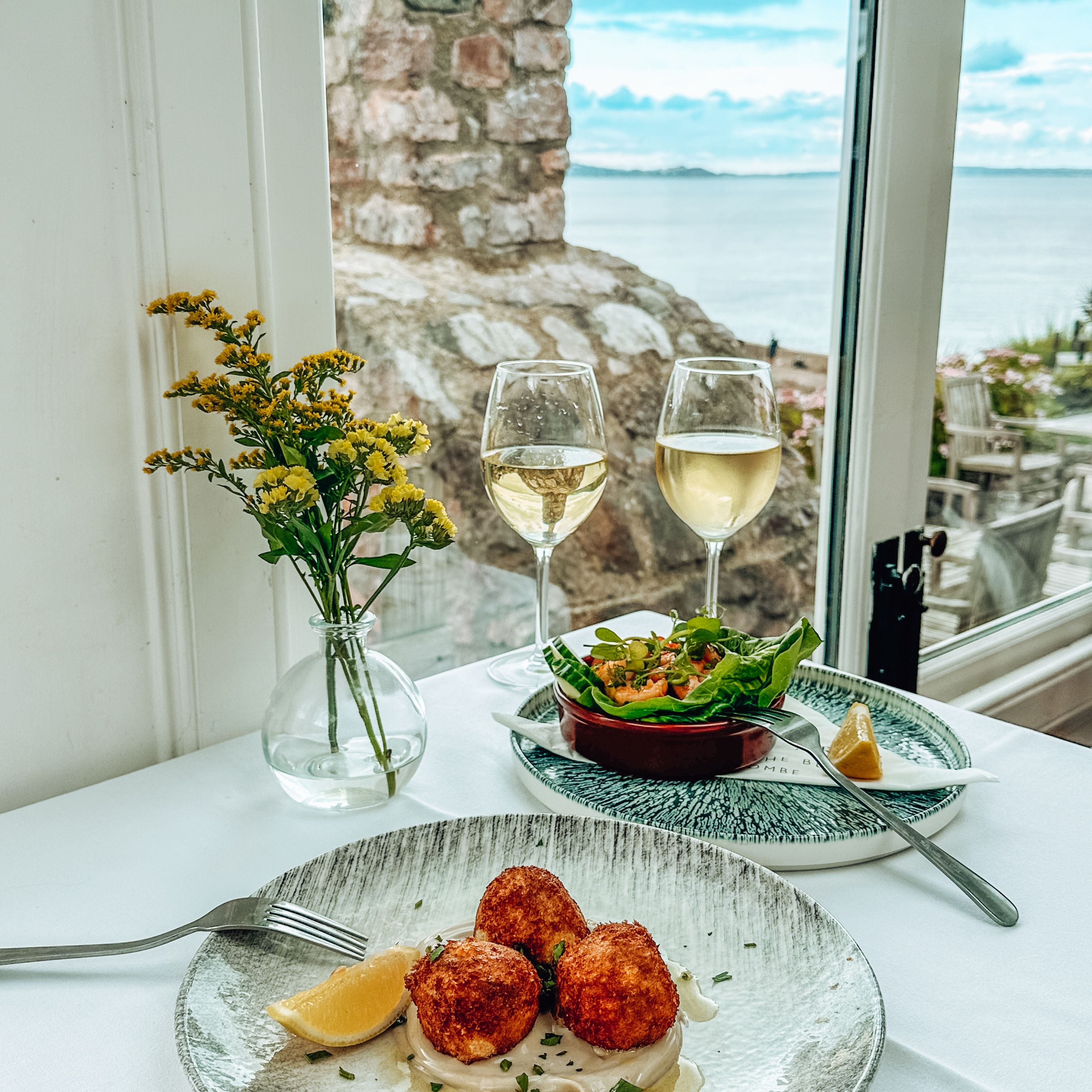 Table set with gourmet seafood appetizers, white wine glasses, and a seaside view.