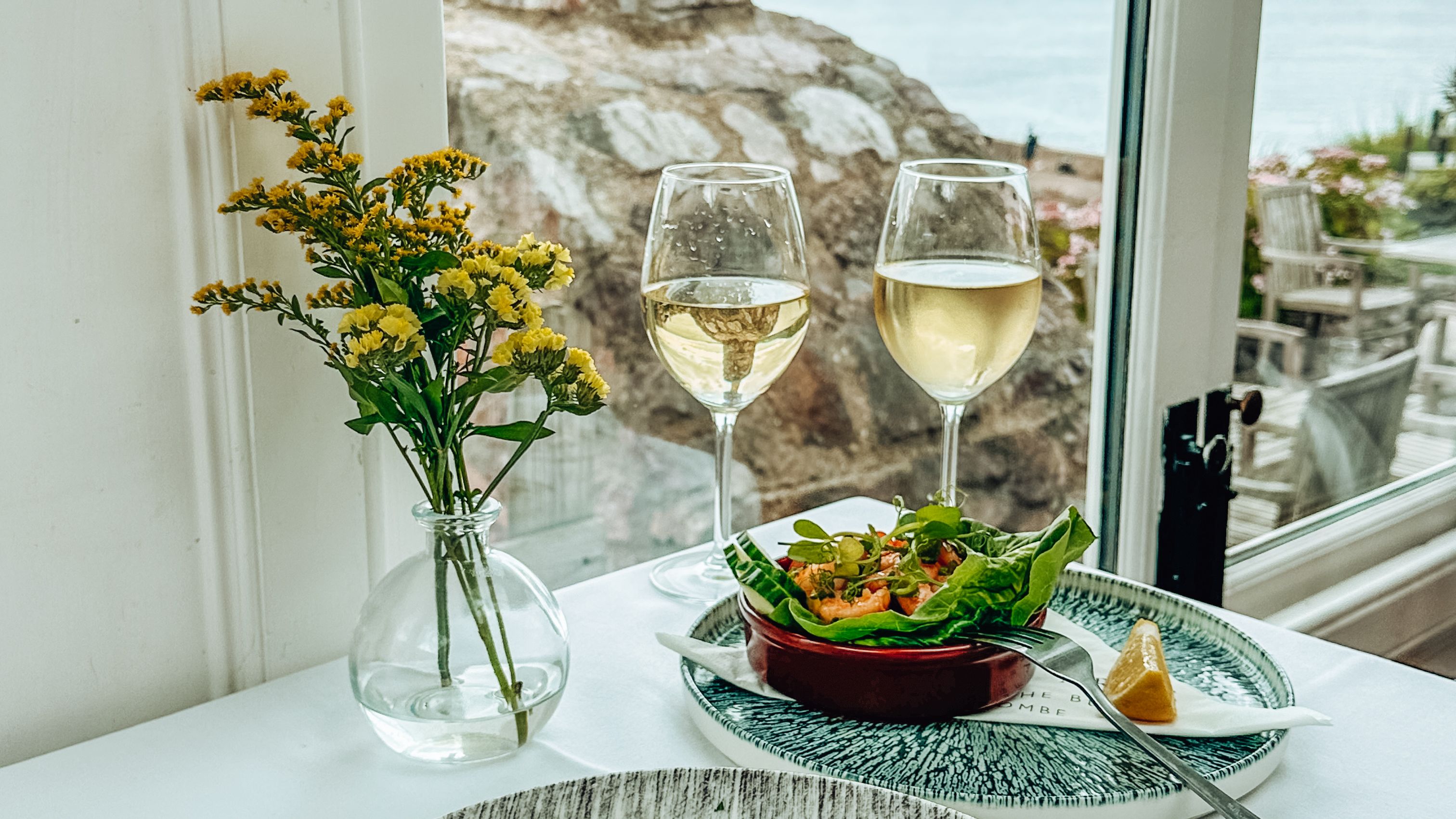 Table set with gourmet seafood appetizers, white wine glasses, and a seaside view.