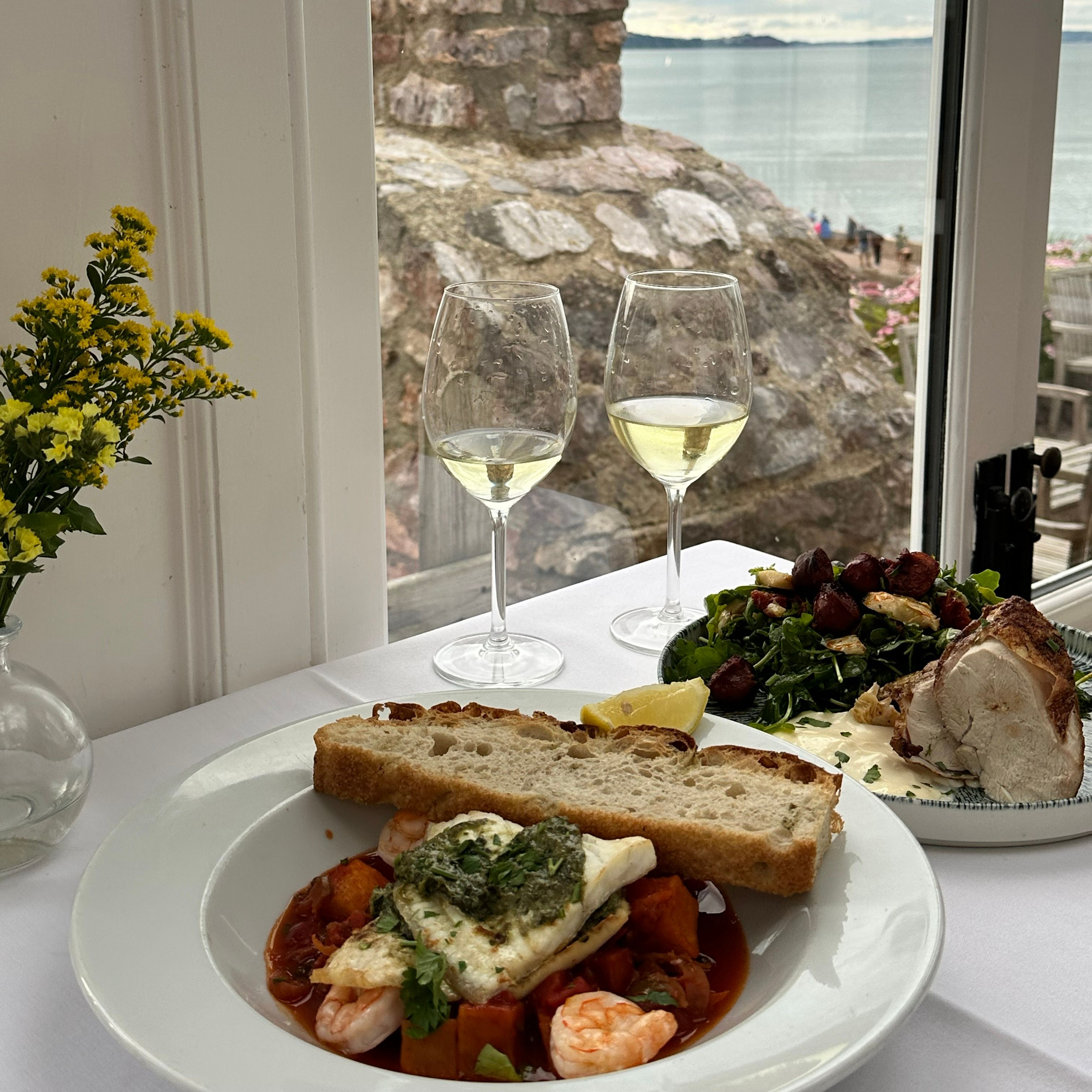 A table set for two by a window, featuring seafood and meat dishes with glasses of white wine overlooking the sea.