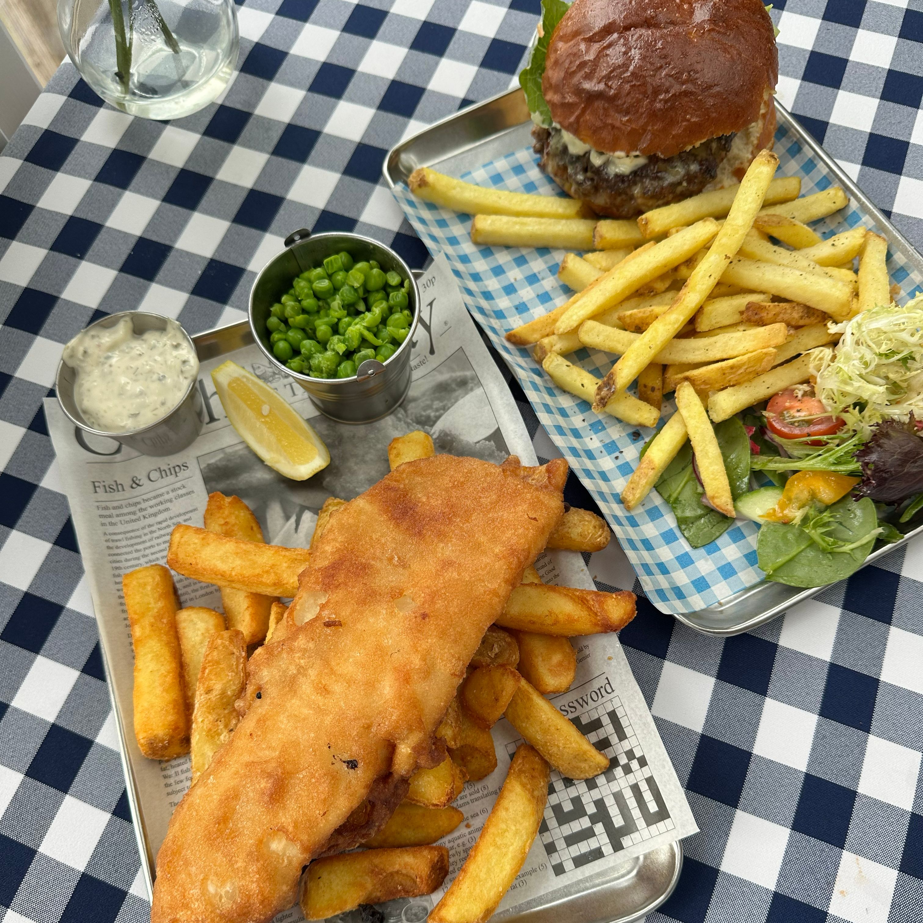 Fish and chips with peas, tartar sauce, and lemon, next to a hamburger with fries and salad on a checkered tablecloth.