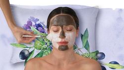 Woman receiving a facial treatment with two types of face masks applied, one dark and one white, while lying on a decorative pillow with botanical illustrations.