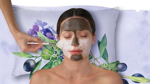 Woman receiving a facial treatment with two types of face masks applied, one dark and one white, while lying on a decorative pillow with botanical illustrations.
