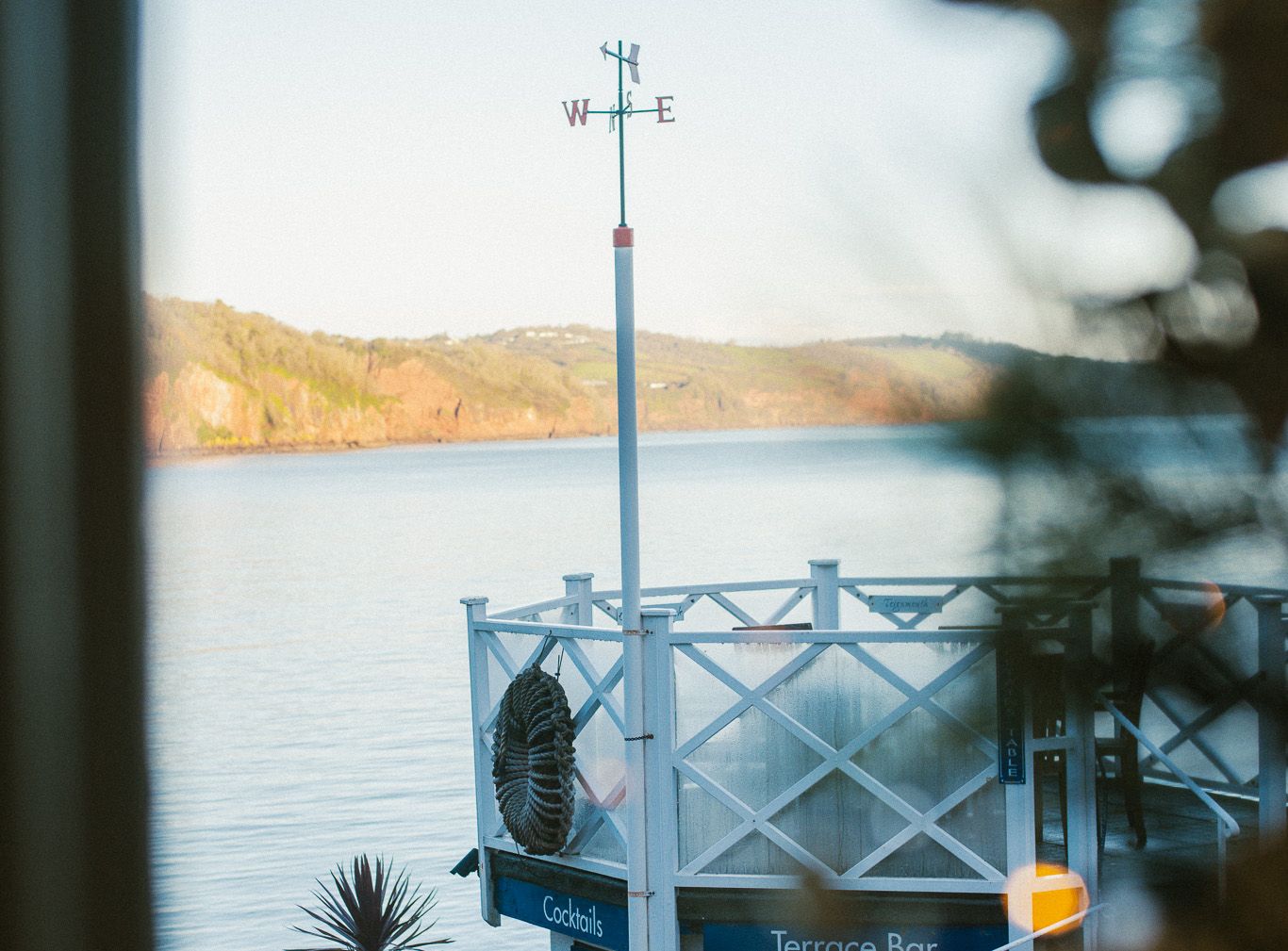 View of a waterfront terrace bar with a weather vane and outdoor seating, overlooking a calm body of water and distant hills.